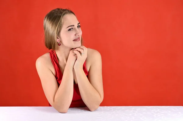 Concepto universal retrato femenino sobre fondo rojo. Foto horizontal de una chica muy sonriente con el pelo largo y un gran maquillaje sentado en una mesa blanca en el estudio . — Foto de Stock