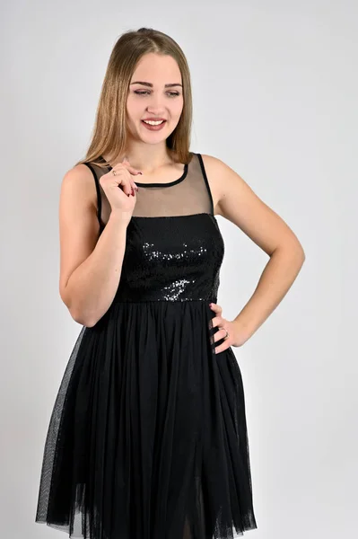Een foto van een mooi lachend meisje met lang haar en uitstekende make-up in een zwarte jurk staat in verschillende poses in de studio. Universeel concept verticale vrouw portret op een witte achtergrond. — Stockfoto