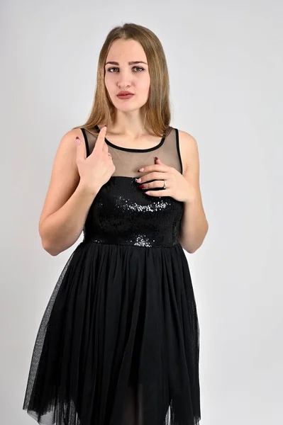 Uma foto de uma menina muito sorridente com cabelo comprido e excelente maquiagem em um vestido preto está de pé em várias poses no estúdio. Universal conceito vertical mulher retrato sobre um fundo branco . — Fotografia de Stock