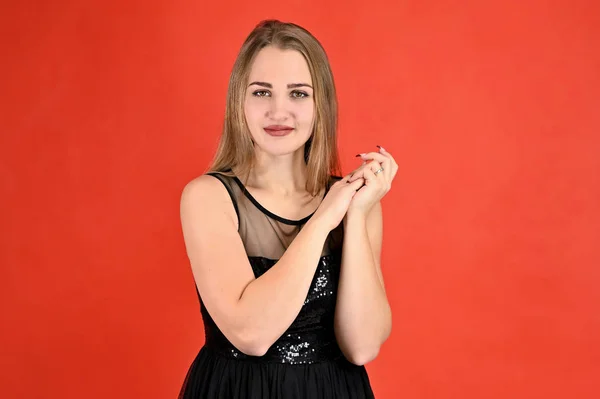 Uma foto de uma menina muito sorridente com cabelo comprido e excelente maquiagem em um vestido preto fica em poses diferentes. Universal conceito horizontal retrato feminino sobre um fundo vermelho . — Fotografia de Stock