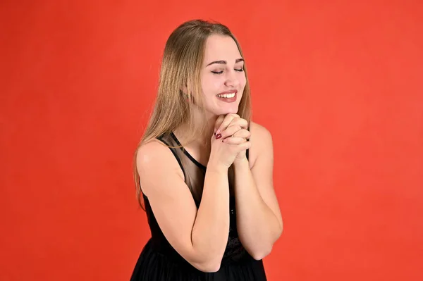 Uma foto de uma menina muito sorridente com cabelo comprido e excelente maquiagem em um vestido preto fica em poses diferentes. Universal conceito horizontal retrato feminino sobre um fundo vermelho . — Fotografia de Stock