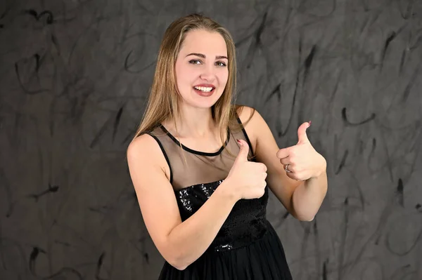 A photo of a pretty smiling girl with long hair and excellent make-up in a black dress stands in different poses. Universal concept of horizontal female portrait on gray alternative background. — 스톡 사진