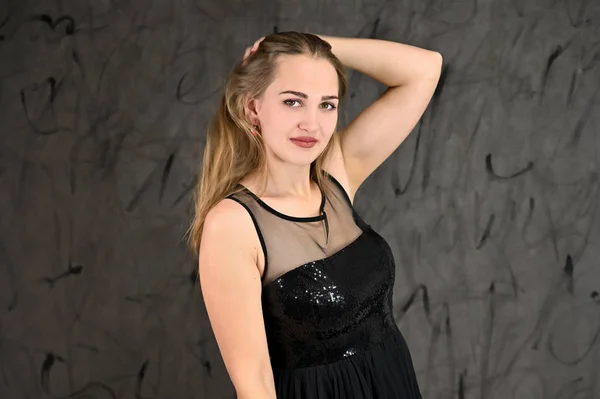 Μια φωτογραφία ενός όμορφου χαμογελαστού κοριτσιού με μακριά μαλλιά και εξαιρετικό μακιγιάζ σε μαύρο φόρεμα στέκεται σε διαφορετικές πόζες. Καθολική έννοια του οριζόντιου γυναικείου πορτραίτου σε γκρι εναλλακτικό φόντο. — Φωτογραφία Αρχείου