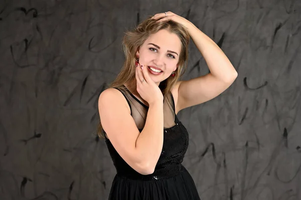 Una foto de una chica muy sonriente con el pelo largo y excelente maquillaje en un vestido negro se encuentra en diferentes poses. Concepto universal de retrato femenino horizontal sobre fondo alternativo gris . — Foto de Stock