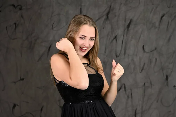 A photo of a pretty smiling girl with long hair and excellent make-up in a black dress stands in different poses. Universal concept of horizontal female portrait on gray alternative background. — Stock Photo, Image