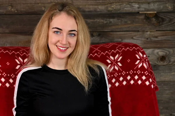 Porträt einer hübschen jungen blonden Frau mit minimalem Make-up in einem Sportdress auf einem hölzernen Hintergrund. sitzt in verschiedenen Posen auf einem roten Sofa gegenüber der Kamera. — Stockfoto