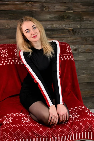 Portret van een mooie jonge blonde vrouw met minimale make-up in een sportjurk op een houten achtergrond. Zit op een rode bank tegenover de camera in verschillende houdingen. — Stockfoto