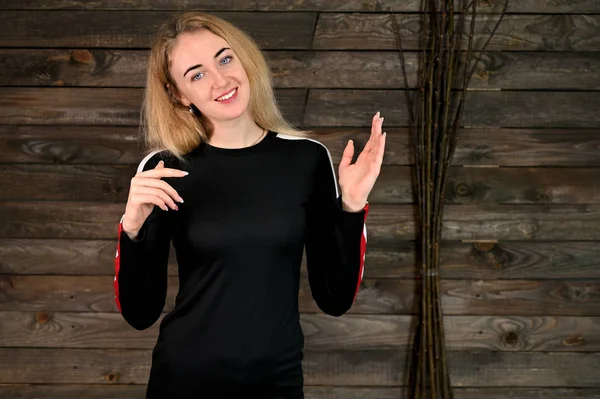 Portrait of a pretty smiling young blonde woman with minimal makeup in a black t-shirt on a wooden background. It stands opposite the camera in various poses. — Stock Photo, Image