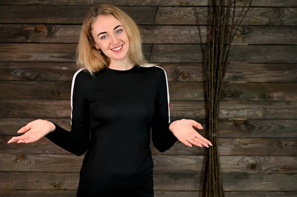 Güzel, gülümseyen sarışın bir kadının portresi. Asgari makyajlı, siyah tişörtlü, tahta arka planda. Kameranın karşısında çeşitli pozlarda duruyor..