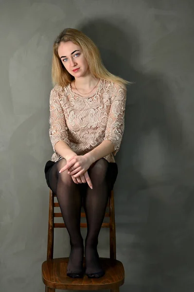 Porträtt av en ganska leende ung blond kvinna med minimal makeup i en beige blus på en grå bakgrund. Sitter på en stol mittemot kameran i alternativa lägen. — Stockfoto