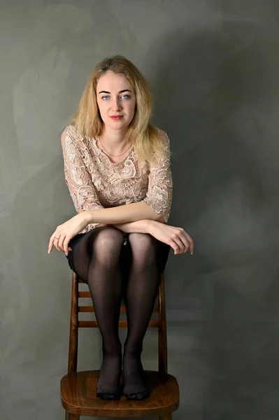 Portret van een mooie glimlachende jonge blonde vrouw met minimale make-up in een beige blouse op een grijze achtergrond. Zit op een stoel tegenover de camera in alternatieve posities. — Stockfoto