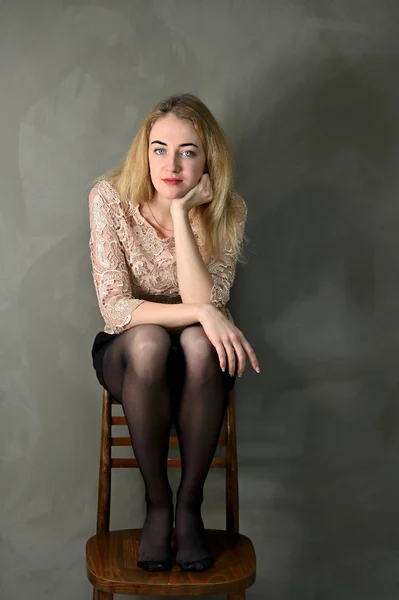 Porträtt av en ganska leende ung blond kvinna med minimal makeup i en beige blus på en grå bakgrund. Sitter på en stol mittemot kameran i alternativa lägen. — Stockfoto