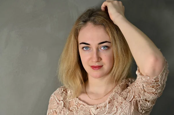 Porträt einer hübschen lächelnden jungen blonden Frau mit minimalem Make-up in einer beigen Bluse auf grauem Hintergrund. er steht der Kamera in verschiedenen Posen direkt gegenüber. — Stockfoto