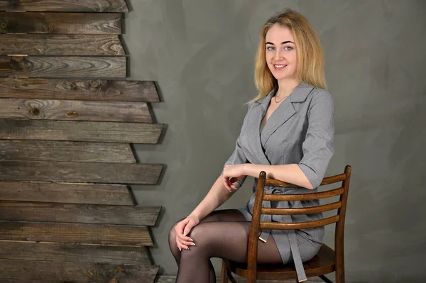Porträtt av en ganska söt leende ung blond affärskvinna med minimal makeup i en grå kostym på en grå bakgrund. Sitter på en stol mittemot kameran i olika poser. — Stockfoto