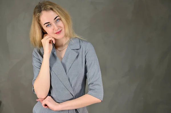 Porträtt av en ganska söt leende ung blond affärskvinna med minimal makeup i en grå kostym på en grå bakgrund. Den står mittemot kameran i olika poser. — Stockfoto