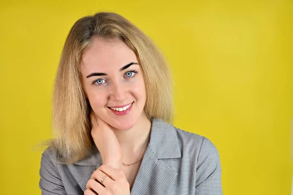 Närbild porträtt av en ganska söt leende ung blond affärskvinna med minimal makeup i en grå kostym på en gul bakgrund. Den står mittemot kameran i olika poser. — Stockfoto