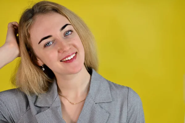 Close-up portrait of a pretty cute smiling young blonde business woman with minimal makeup in a gray suit on a yellow background. It stands directly opposite the camera in various poses. — Stock Photo, Image