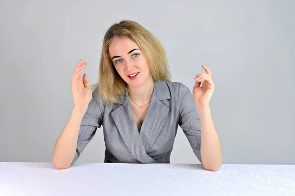 Retrato de una linda joven mujer de negocios rubia sonriente con un maquillaje mínimo en un traje gris sobre un fondo blanco. Modelo Se sienta en una mesa justo enfrente de la cámara en varias poses . — Foto de Stock