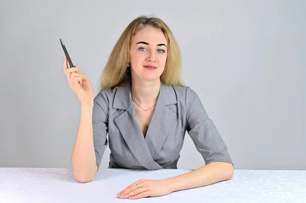 Retrato de una linda joven mujer de negocios rubia sonriente con un maquillaje mínimo en un traje gris sobre un fondo blanco. Modelo Se sienta en una mesa justo enfrente de la cámara en varias poses . — Foto de Stock