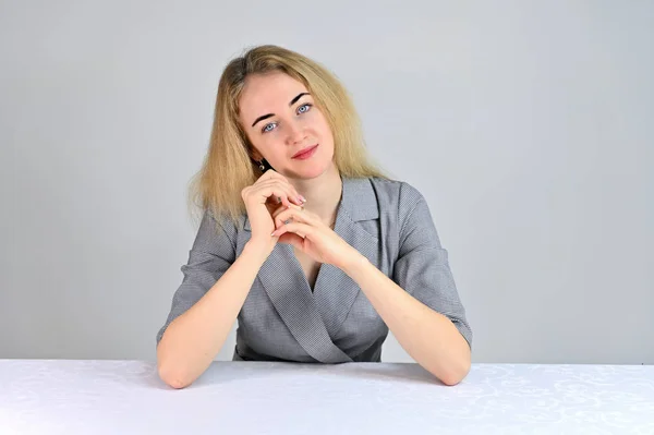 Modell Sitter vid ett bord mittemot kameran i olika poser. Porträtt av en ganska söt leende ung blond affärskvinna med minimal makeup i en grå kostym på en vit bakgrund. — Stockfoto