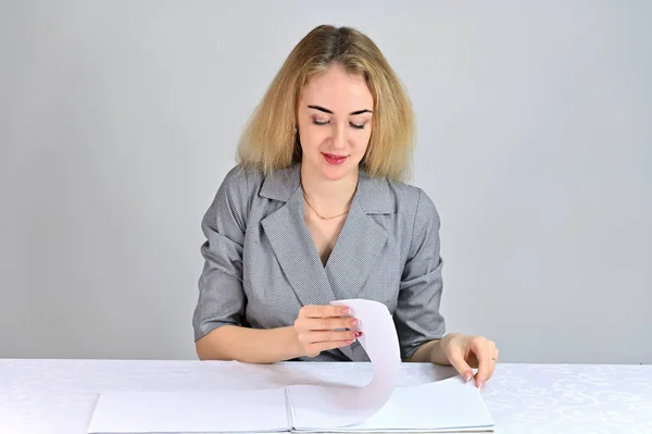 Modello Si siede a un tavolo direttamente di fronte alla fotocamera in varie pose. Ritratto di una giovane donna d'affari bionda piuttosto carina e sorridente con un trucco minimale in un abito grigio su uno sfondo bianco . — Foto Stock