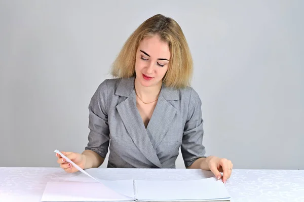 Modelo Se sienta en una mesa directamente enfrente de la cámara en varias poses. Retrato de una linda joven mujer de negocios rubia sonriente con un maquillaje mínimo en un traje gris sobre un fondo blanco . — Foto de Stock