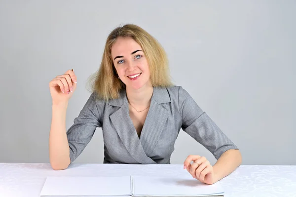 Modelo Se sienta en una mesa directamente enfrente de la cámara en varias poses. Retrato de una linda joven mujer de negocios rubia sonriente con un maquillaje mínimo en un traje gris sobre un fondo blanco . — Foto de Stock