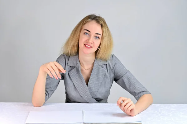 Model Sits at a table directly opposite the camera in various poses. Portrait of a pretty cute smiling young blonde business woman with minimal makeup in a gray suit on a white background. — Stock Photo, Image