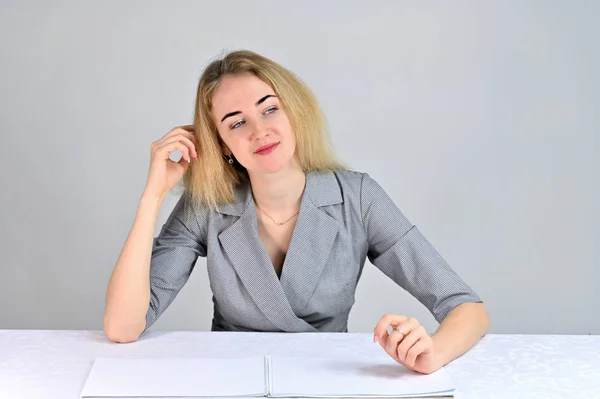 Model Sits at a table directly opposite the camera in various poses. Portrait of a pretty cute smiling young blonde business woman with minimal makeup in a gray suit on a white background. — Stock Photo, Image