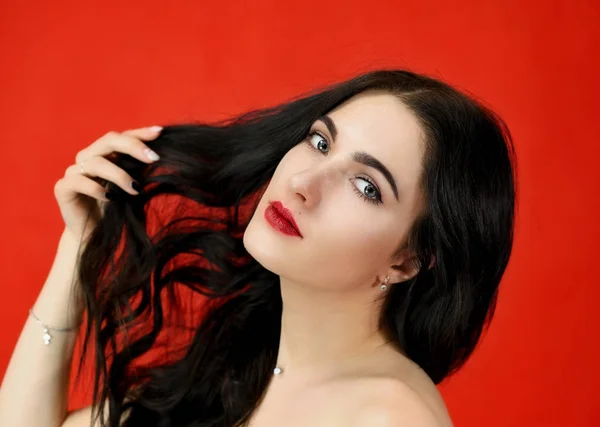 Le concept de longs et beaux cheveux. Gros plan Portrait d'une jolie fille modèle brune avec un excellent maquillage et des cheveux luxueux sur fond rouge en studio . — Photo