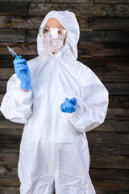 Yanmış bir evin ahşap duvarına karşı beyaz bir tıbbi giysi giymiş bir doktorun fotoğrafı. Elinde şırıngayla kameranın önünde çeşitli pozlarda duruyor..