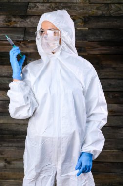 Yanmış bir evin ahşap duvarına karşı beyaz bir tıbbi giysi giymiş bir doktorun fotoğrafı. Elinde şırıngayla kameranın önünde çeşitli pozlarda duruyor..