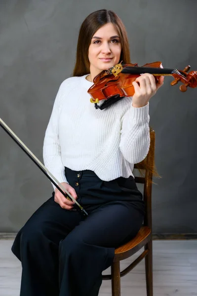 Portrait of a pretty brunette musician girl in a white jacket on a gray background sits on a chair with a violin in her hands — Stock Photo, Image