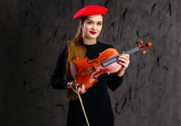 Retrato de uma menina músico morena bonita em um vestido preto e boina vermelha em um fundo cinza toca violino — Fotografia de Stock