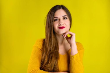 Sarı arka planda sarı bir süveter giyen uzun saçlı güzel bir esmer kızın portresi gülümsüyor ve olumlu duygular gösteriyor.
