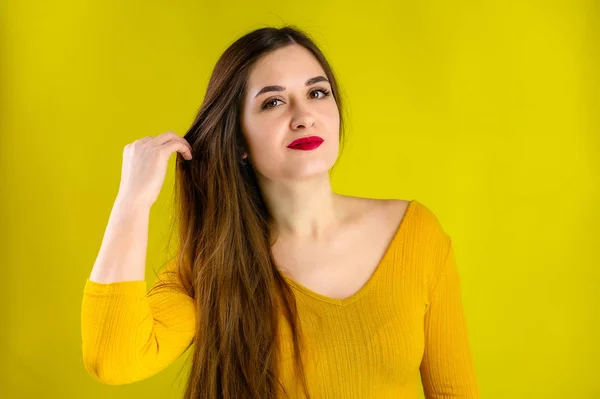 Portrét krásné brunetky s dlouhými vlasy s úsměvem ve žlutém svetru na žlutém pozadí úsměvy a ukazuje pozitivní emoce — Stock fotografie