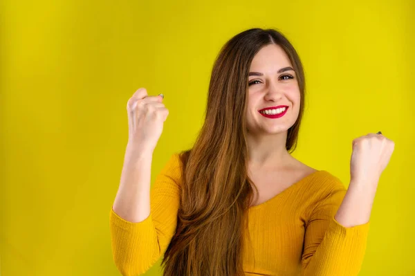 Buena chica morena con el pelo largo con una sonrisa en una chaqueta amarilla se regocija sobre un fondo amarillo, sonríe y muestra emociones positivas — Foto de Stock