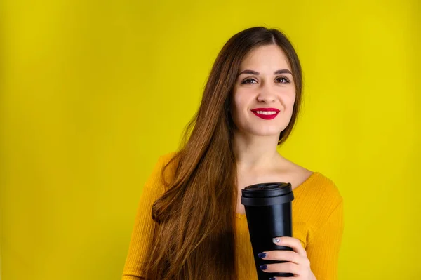 Menina morena agradável com cabelos longos com um sorriso com uma xícara de café em uma camisola amarela é feliz em um fundo amarelo sorrindo e mostrando emoções positivas — Fotografia de Stock