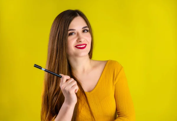 Menina morena agradável com cabelos longos com um sorriso com uma caneta em suas mãos em uma camisola amarela é feliz sobre um fundo amarelo sorrindo e mostrando emoções positivas — Fotografia de Stock