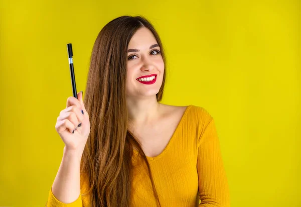 Menina morena agradável com cabelos longos com um sorriso com uma caneta em suas mãos em uma camisola amarela é feliz sobre um fundo amarelo sorrindo e mostrando emoções positivas — Fotografia de Stock