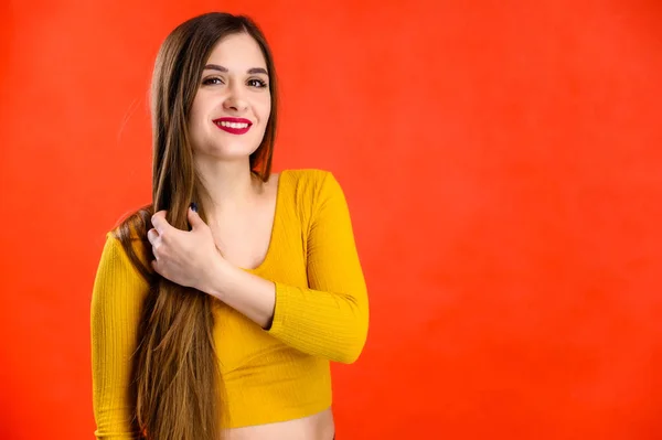 Buena chica morena con el pelo largo con una sonrisa en una sudadera amarilla se regocija sobre un fondo rojo sonríe y muestra emociones positivas — Foto de Stock