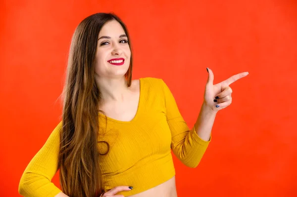 Menina morena agradável com cabelos longos com um sorriso em uma camisola amarela se alegra em um fundo vermelho sorri e mostra emoções positivas — Fotografia de Stock