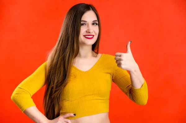 Menina morena agradável com cabelos longos com um sorriso em uma camisola amarela se alegra em um fundo vermelho sorri e mostra emoções positivas — Fotografia de Stock