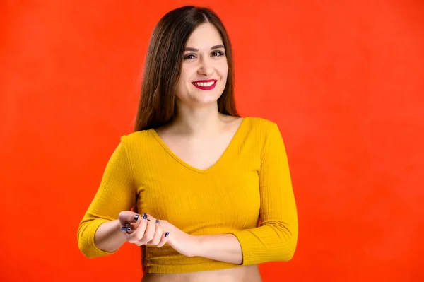 Menina morena agradável com cabelos longos com um sorriso em uma camisola amarela se alegra em um fundo vermelho sorri e mostra emoções positivas — Fotografia de Stock