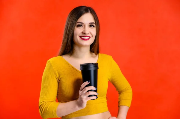 Menina morena agradável com cabelos longos com um sorriso com uma xícara de café em suas mãos em uma camisola amarela é feliz sobre um fundo vermelho sorrindo e mostrando emoções positivas — Fotografia de Stock