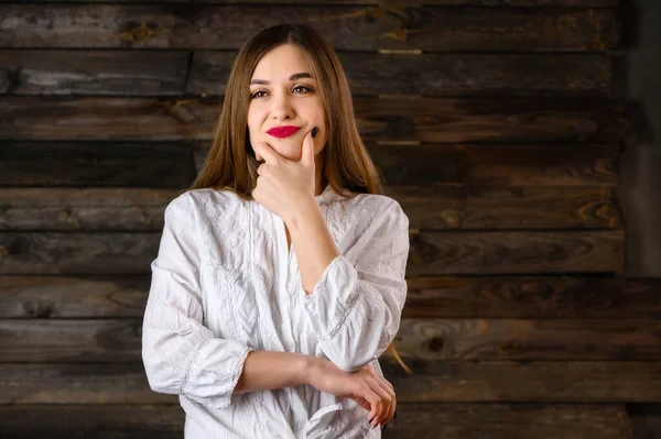 Buena chica morena con el pelo largo con una sonrisa en una camisa blanca se comunica sobre un fondo de madera, sonríe y muestra emociones positivas — Foto de Stock