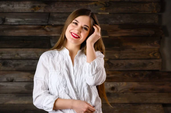 Buena chica morena con el pelo largo con una sonrisa en una camisa blanca se comunica sobre un fondo de madera, sonríe y muestra emociones positivas — Foto de Stock