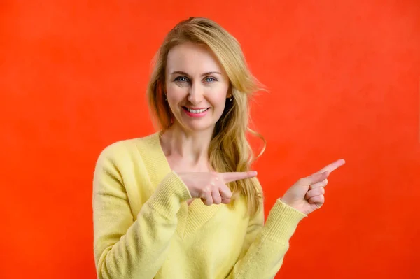 Фото 40-летней блондинки в жёлтом свитере, показывающей пальцы на красном фоне — стоковое фото