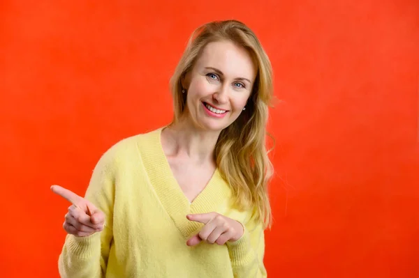 Mulher loira de 40 anos em uma camisola amarela mostra os dedos em um fundo vermelho — Fotografia de Stock