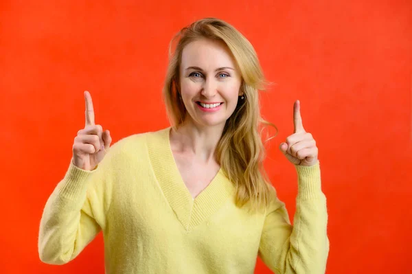 Loira bonita 40 anos de idade em uma camisola amarela mostra seus dedos com alegria em um fundo vermelho — Fotografia de Stock